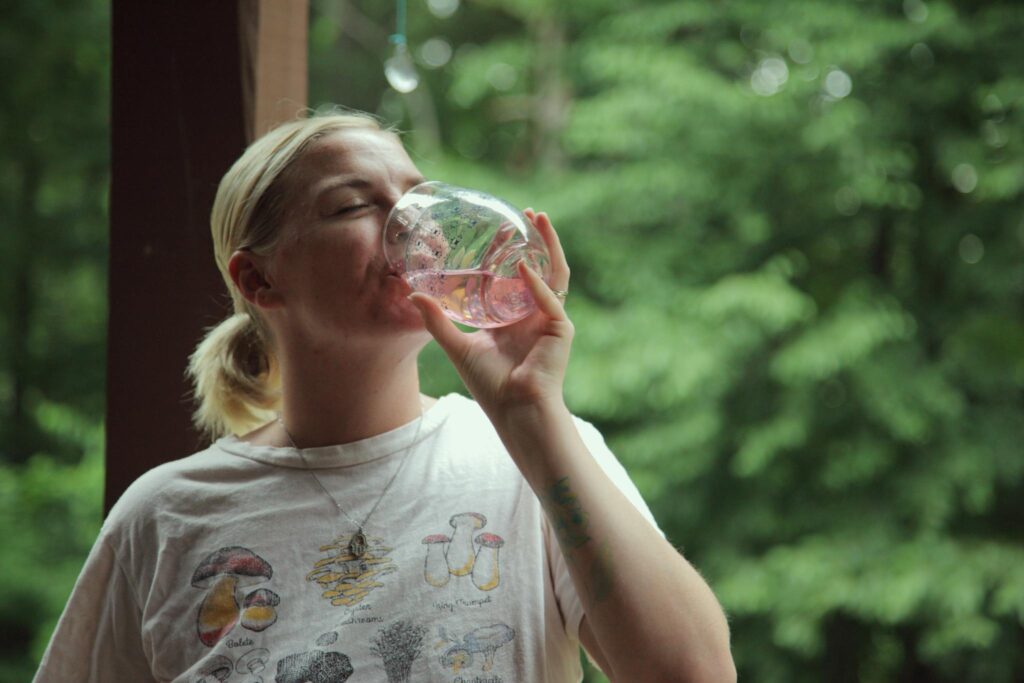 A woman drinks water from a glass with a peaceful forest background, enjoying nature.